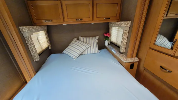 Cozy RV rental bedroom with light blue sheets, striped pillows, overhead cabinets, and window blinds.. Thor Freedom Elite 2017