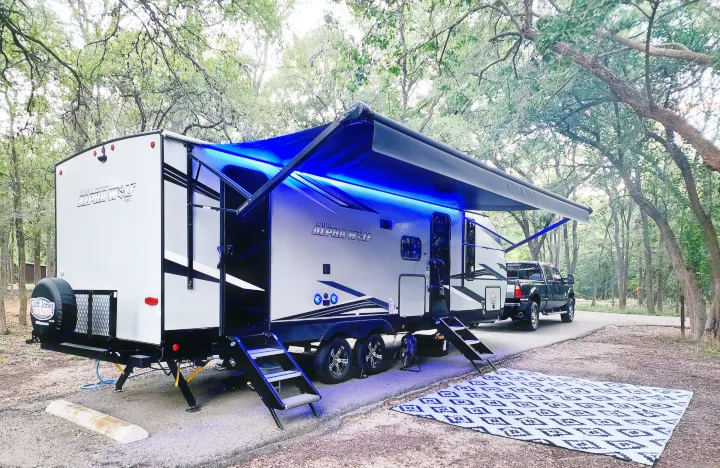 Modern Alpha Wolf RV rental at a wooded campsite, featuring an illuminated blue awning and outdoor mat.. Forest River Cherokee Alpha Wolf 2022
