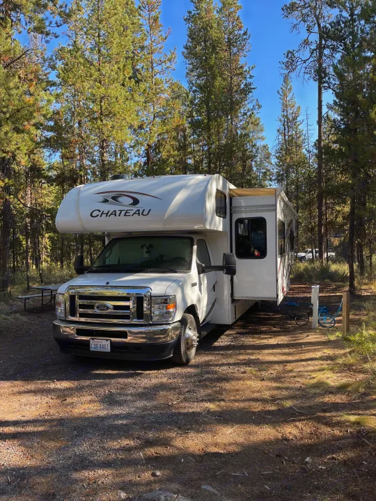 A white Chateau rv rental is set up at a forest campsite. Its slide-out is extended, offering comfortable camping among tall pine trees.. Thor Chateau 2022