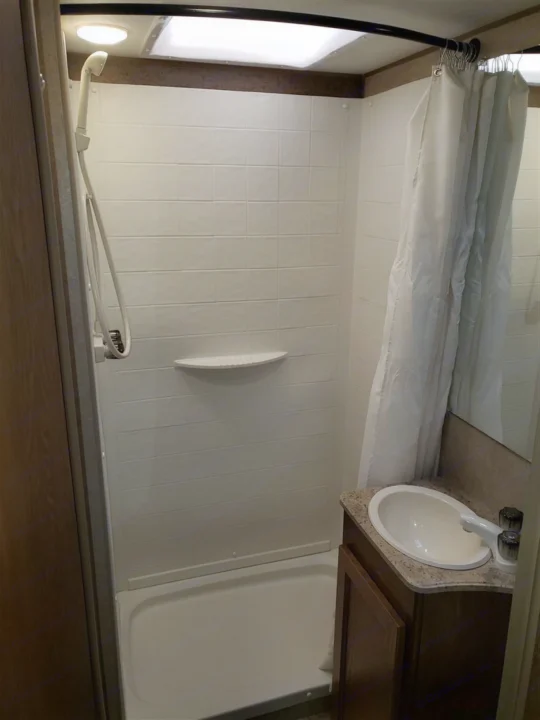 This compact RV rental bathroom includes a white shower stall with a handheld showerhead, a small sink, and a mirror.. Forest River Coachmen Leprechaun 2017