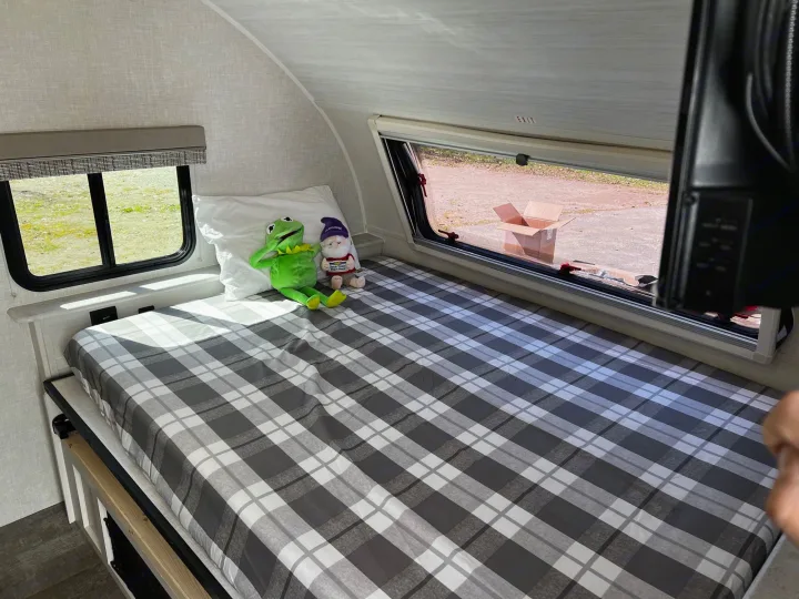Relaxing RV rental interior: a comfortable plaid bed with frog and gnome plushies, sunlit by a large window overlooking outside.. Forest River R POD 193  H.R. EDITION 2022