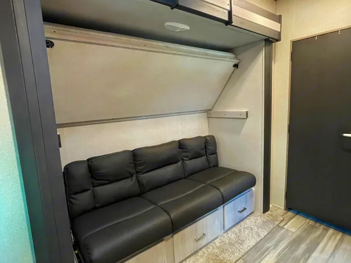 This RV rental interior shows a black sofa with storage, a fold-down bunk bed, and a dark door.. Keystone RV Sprinter 2021