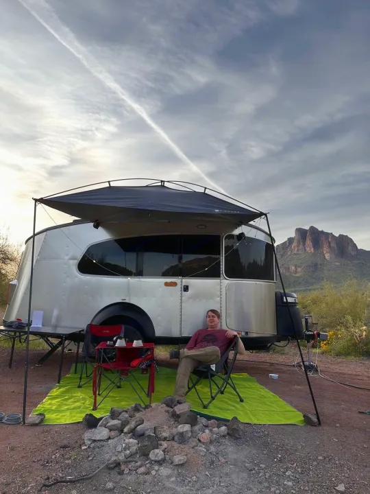 Enjoy Arizona's beauty with an RV rental. A person relaxes by their travel trailer at a desert campsite under dusk skies.. Airstream Basecamp 2021
