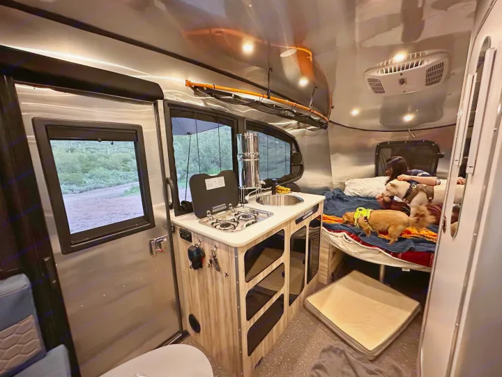 RV rental interior: A compact kitchen, bed with a person & two dogs, and suspended ceiling storage.. Airstream Basecamp 2021