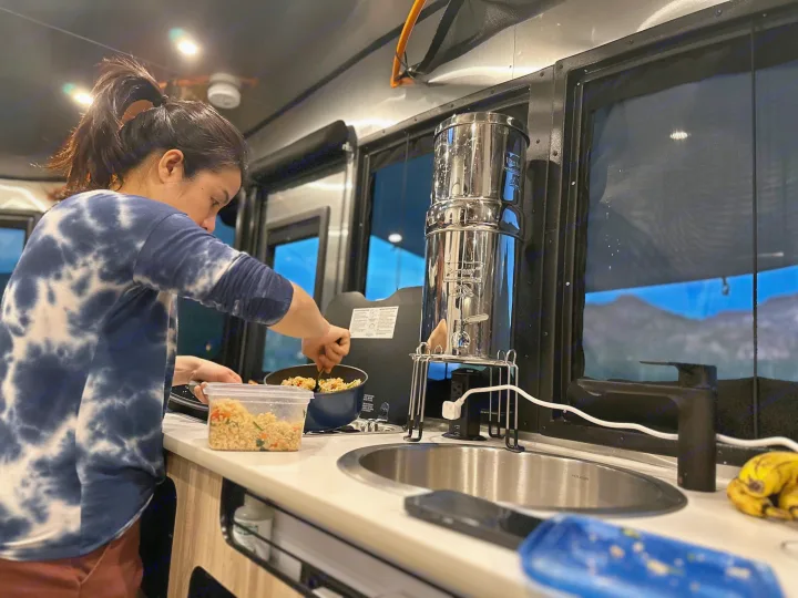 Person cooks in an RV rental kitchen, featuring a portable water filter. Mountain views visible through the window.. Airstream Basecamp 2021