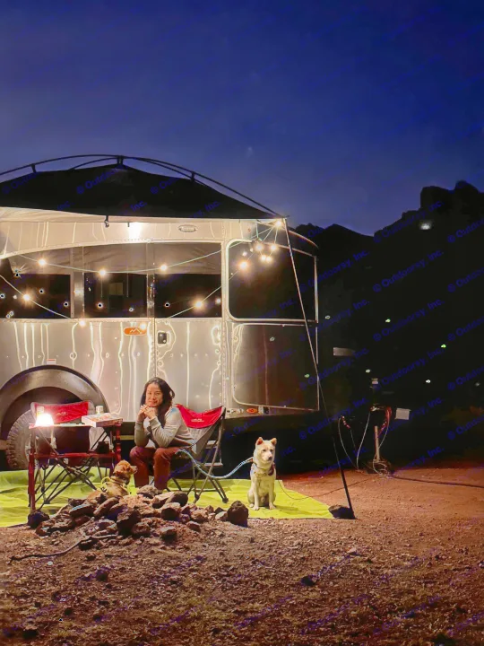 Enjoying a cozy night at an RV rental campsite. A person, two dogs, and string lights illuminate the silver travel trailer.. Airstream Basecamp 2021
