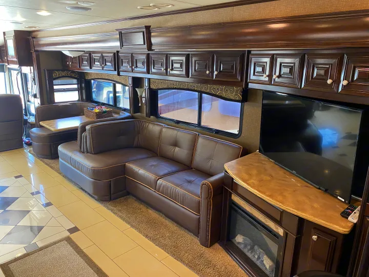 This spacious RV rental interior shows a brown leather sectional, dining booth, TV, and fireplace. Overhead cabinets line the walls.. Thor Motor Coach Tuscany 2015