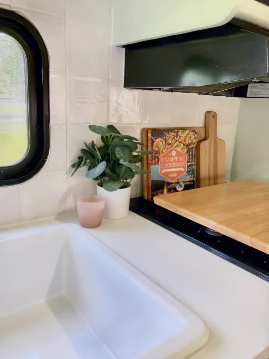 Bright RV rental kitchen with white sink, tiled backsplash, window, plant, and 'Campfire Cookbook' on the counter.. Forest River Other 2013