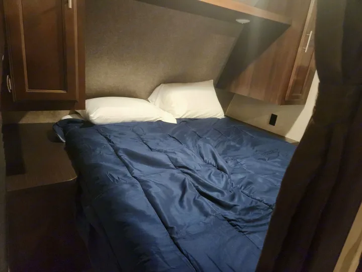 A cozy RV rental bedroom features a bed with a dark blue comforter, white pillows, and wood cabinetry.. Jayco Jay Flight SLX 2019