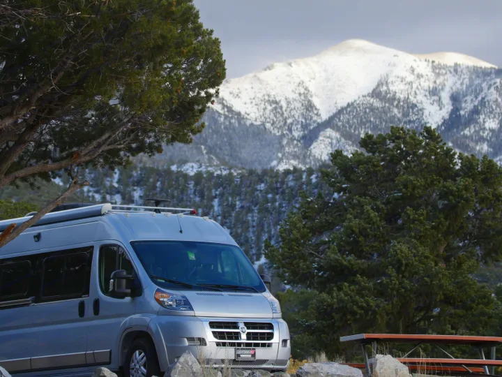 Silver RV rental camper van parked near large trees with snow-capped mountains in the background, offering a winter travel experience.. Winnebago Travato 2018