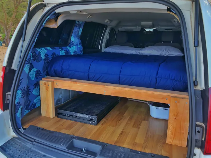 This RV rental shows a cozy van conversion: a bed with blue bedding, under-bed storage, & tropical curtains.. Dodge Grand Caravan 2010