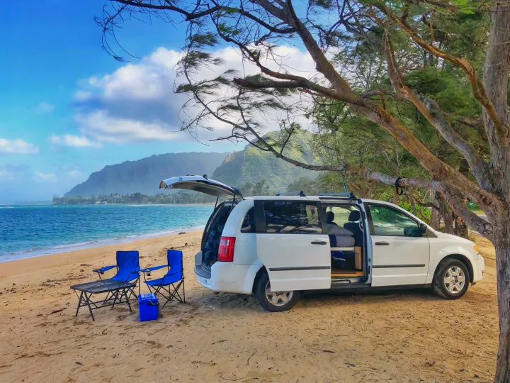 Coastal RV rental adventure: white minivan parked on sandy beach with chairs, overlooking the ocean & mountains.. Dodge Grand Caravan 2010
