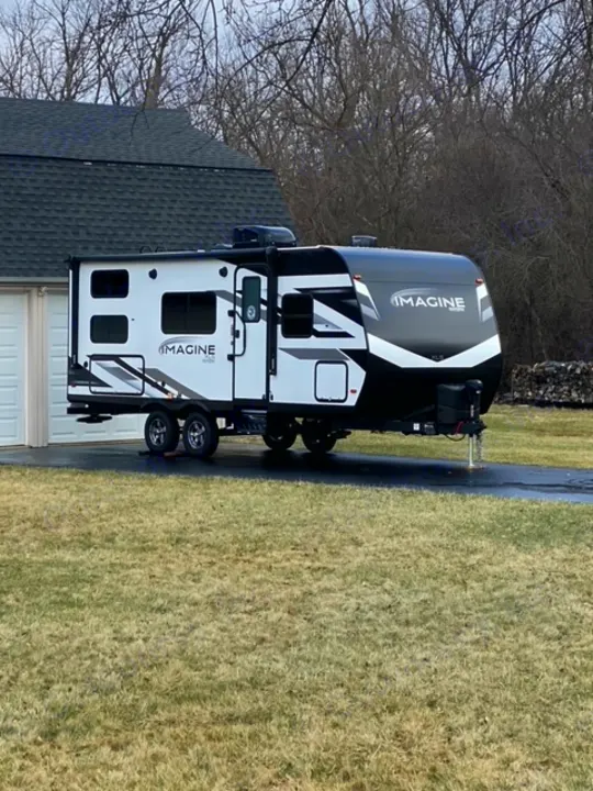 A stylish Grand Design Imagine travel trailer is parked, ready for an rv rental, with a house and grass in view.. Grand Design 21BHE 2023