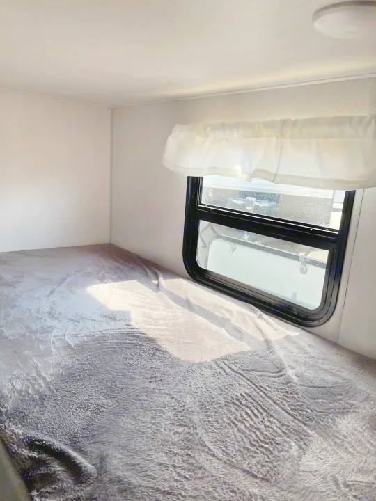 Cozy RV rental sleeping area features a plush gray bed under a window with a sheer white valance, illuminated by sunlight.. Avenger LT 2022