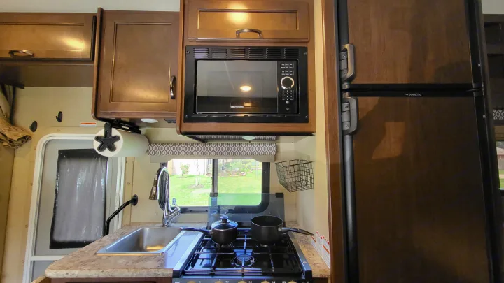 Modern RV rental kitchen with dark wood cabinets, microwave, stove, sink, and Dometic fridge.. Thor 22HE 2019