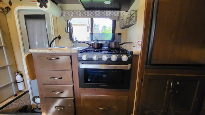 A compact RV kitchen with a stainless steel sink, black stove/oven unit, and wooden drawers. Ideal for an RV rental.. Thor 22HE 2019
