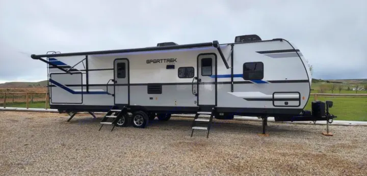 Spacious white & grey RV rental with extended awning parked on gravel. Perfect for travel and camping adventures.. Venture Sporttrek 2022