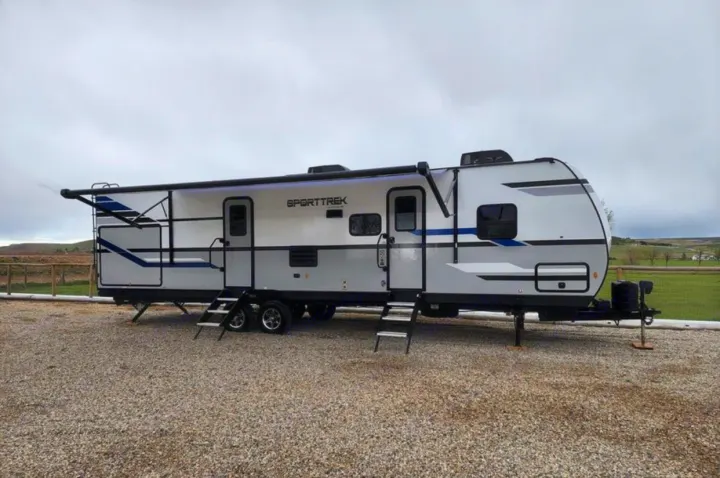 This modern white and gray SportTrek travel trailer, an ideal RV rental, is parked on a gravel lot.. Venture Sporttrek 2022