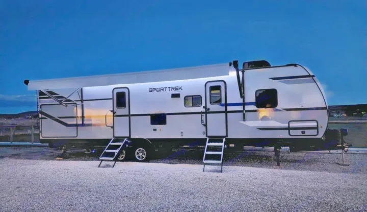 Brightly lit SportTrek RV rental parked on gravel at night, showing its extended awning and two entry doors.. Venture Sporttrek 2022