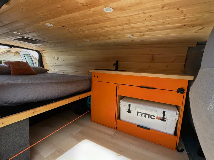 This RV rental showcases a custom interior with a bed, wood paneling, and an orange kitchen cabinet storing an RTIC cooler.. Ford Extended E350 2011