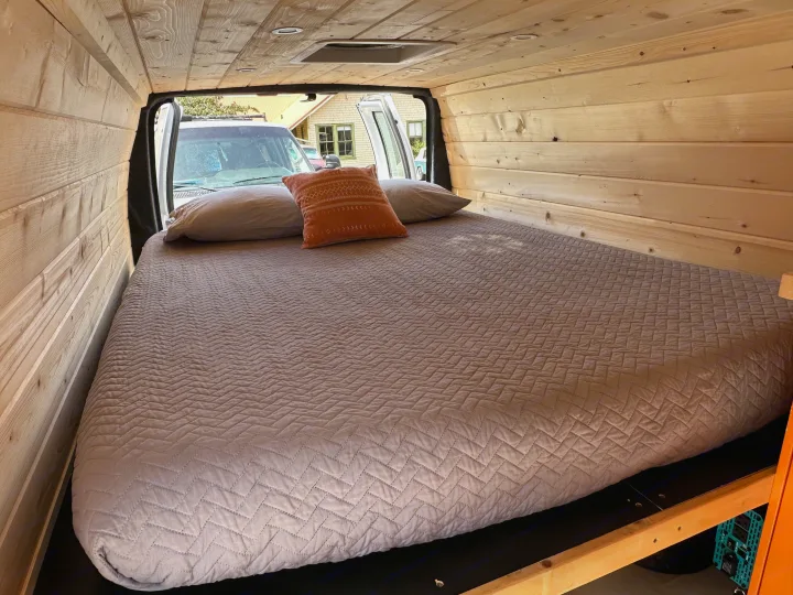 Custom wood paneled RV rental interior with a large comfortable bed and pillows. Rear doors are open, showing the exterior.. Ford Extended E350 2011