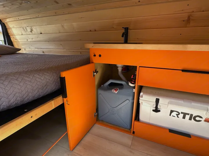 Interior of an RV rental: features a cozy bed, compact kitchen with a sink, water tank, and an RTIC cooler.. Ford Extended E350 2011