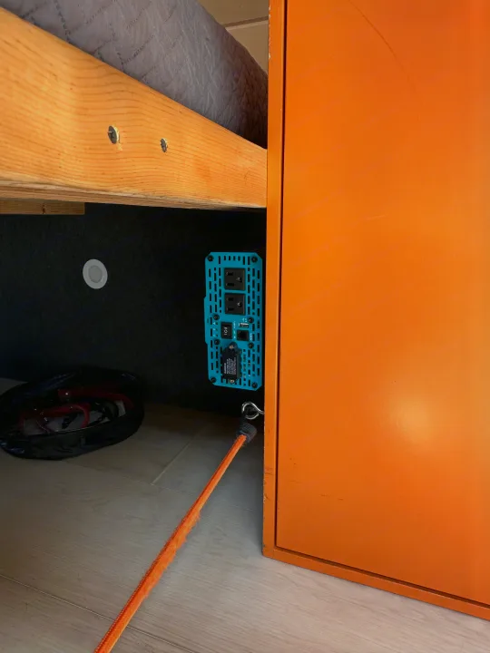 Inside an RV rental: a blue power inverter, light, and cable bag under a wooden bed frame, beside an orange cabinet.. Ford Extended E350 2011