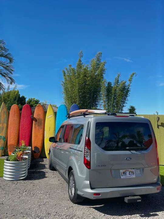 Explore Hawaii with this RV rental: A grey van with surfboards on its roof, parked near a vibrant display of colorful surfboards.. Ford Transit Connect Wagon 2022