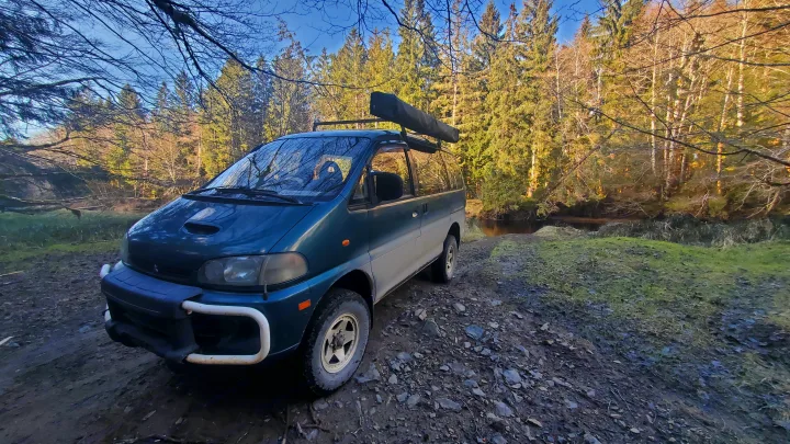 A teal Mitsubishi Delica adventure van, perfect for RV rental, parked by a forest river on a muddy track under a blue sky.. MITSUBISHI Delica 1997