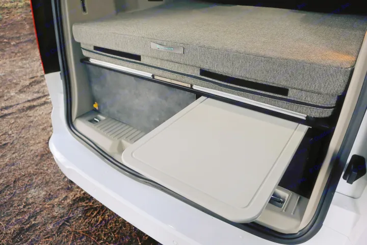 Inside an RV rental: a compact, modular bed with "Caravan Master" branding, and a grey pull-out table extended from beneath.. Ford Transit Connect Wagon 2022