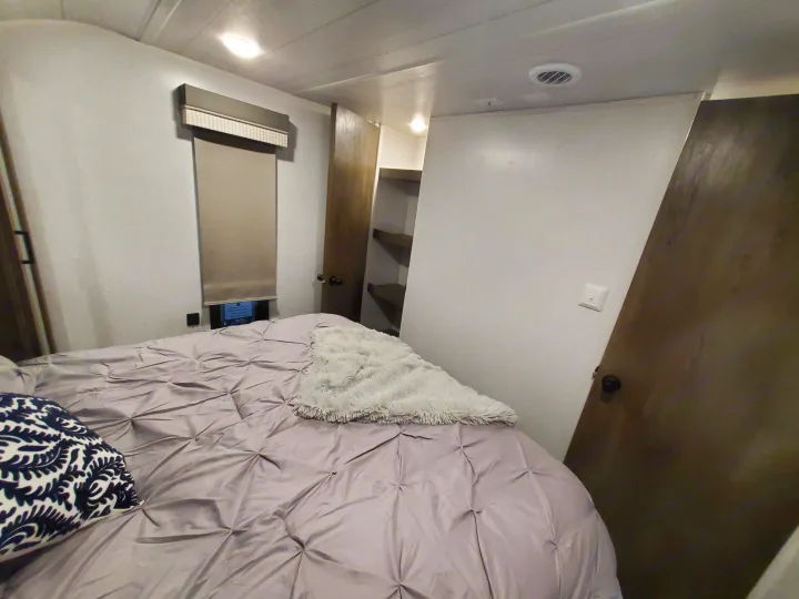 A bright RV rental bedroom features a bed with a light purple comforter, window shade, and open closet shelves.. Forest River Salem 2022