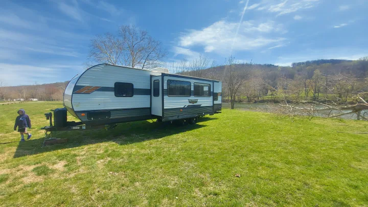 RV rental set up on a grassy riverside spot, offering outdoor camping views with a child nearby.. Forest River Wildwood 2020
