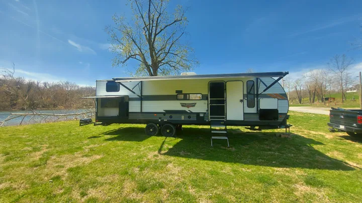 Sunny day RV rental: Wildwood travel trailer parked by a river, ready for camping fun.. Forest River Wildwood 2020