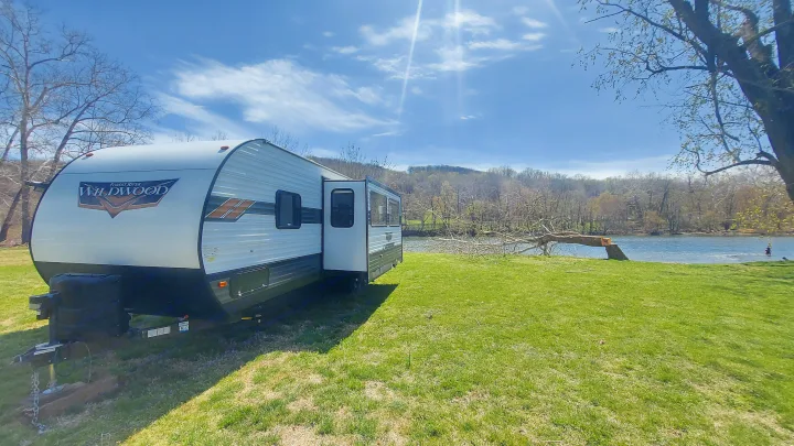 This Forest River Wildwood RV rental is perfectly set up on green grass beside a scenic river on a sunny day.. Forest River Wildwood 2020