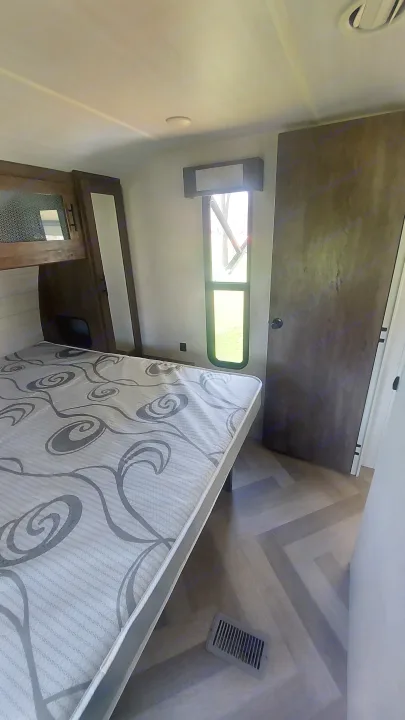 This RV rental features a cozy bedroom with a patterned mattress, wood-look floor, and a long window.. Forest River Wildwood 2020