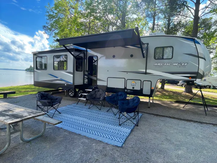 This RV rental offers a perfect lakeside camping experience. A large fifth-wheel with an awning, outdoor rug, and chairs by a scenic lake.. Forest River Cherokee Arctic Wolf 2022