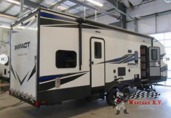 This white & blue toy hauler RV for rv rental features an extended awning & entry steps, parked indoors.. Keystone Impact 2019