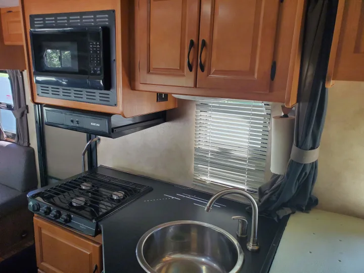 An RV rental kitchen showing a microwave, gas stove, stainless steel sink, wooden cabinets, and a window with blinds.. Thor Majestic 2014