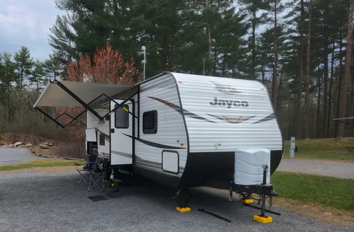 A white Jayco RV rental parked on a gravel campsite, with its awning extended and chairs set up near trees.. Jayco Jay Flight SLX 2019