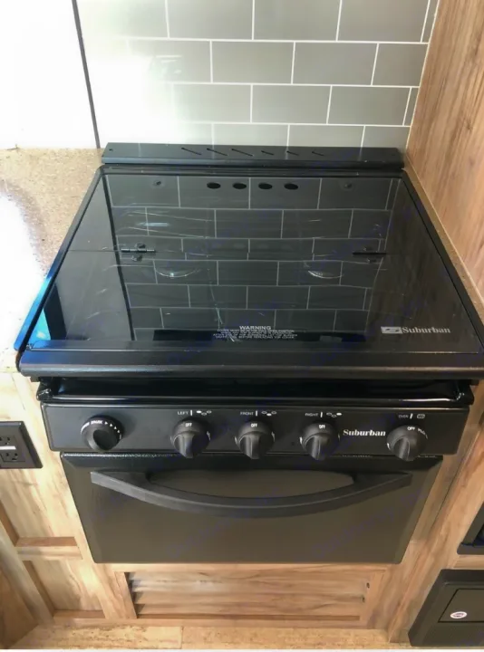 RV rental kitchen with black Suburban gas range and oven. Glass stovetop cover reflects grey backsplash.. Palomino 240bhs 2018