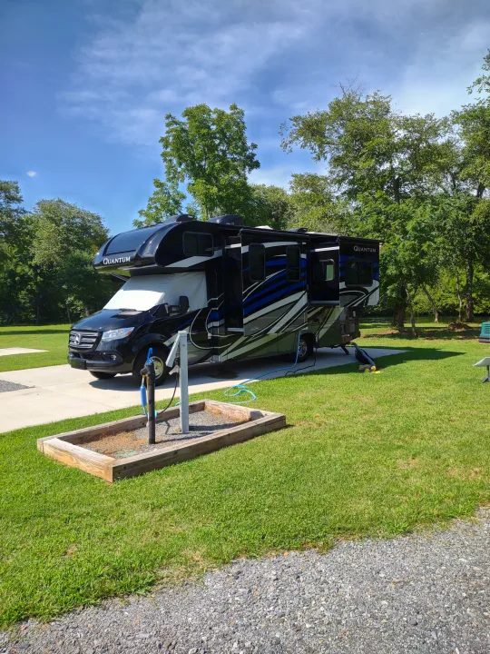 Black and blue Quantum RV rental parked at a campsite with utility hookups, green grass, and trees under a blue sky.. Thor Quantum 2021