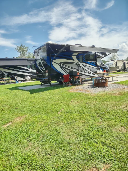 An RV rental, a blue and white Quantum motorhome, parked at a sunny campsite with a fire pit, chairs, and grassy area.. Thor Quantum 2021