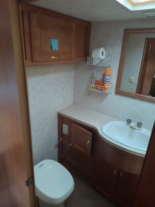 Inside an RV rental: a neat bathroom with toilet, sink, wood cabinets, and wire shelves holding Lysol & toilet paper.. Damon Corporation Daybreak 2001
