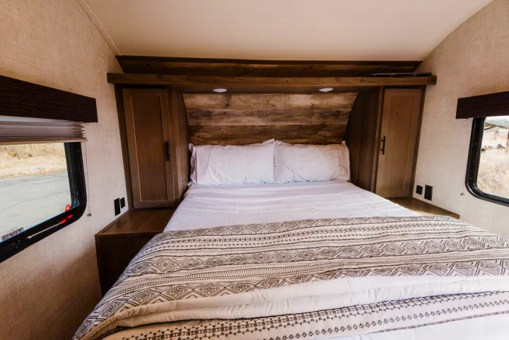 RV rental bedroom features a comfy queen bed with white linens & patterned throw, wood headboard, storage, and side windows.. Gulf Stream Ameri-Lite 2021