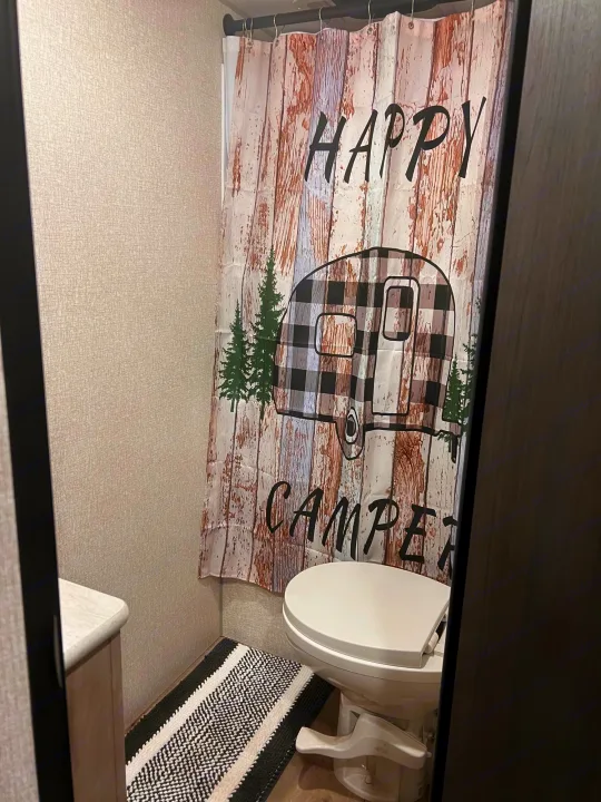 Compact RV rental bathroom features a "Happy Camper" shower curtain, toilet, and striped rug.. Escape e231bh 2022