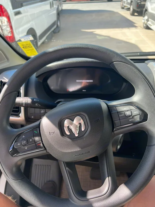 POV from an RV rental: Ram ProMaster steering wheel with controls, dashboard display, and a parked white van visible outside.. Thor Tellaro 2023