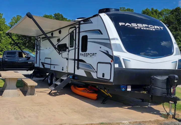 White Passport GT Series RV rental parked at a sunny campsite with an extended awning, pickup truck, concrete picnic table, and kayak.. Keystone Passport 2022