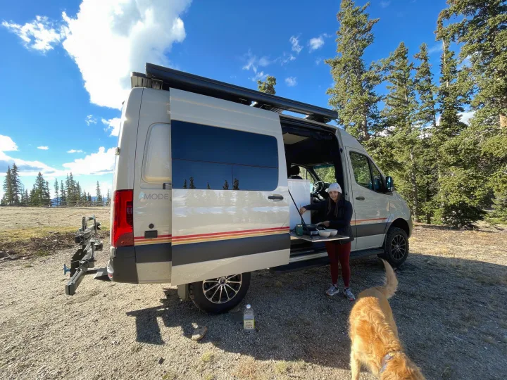 RV rental freedom: Woman prepares meal from her Mode camper van, dog nearby, amidst scenic mountain trees.. StoryTeller Overland Classic Mode 4X4 2022