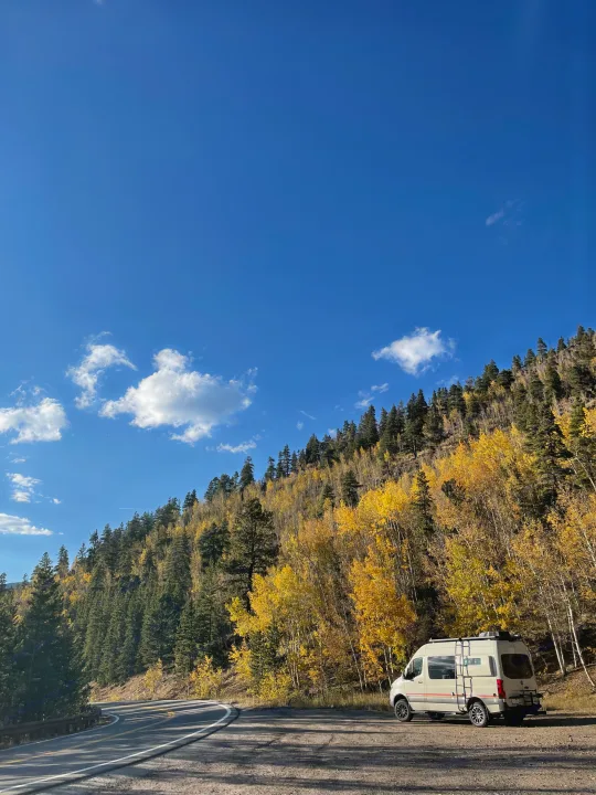 An RV rental is parked on a scenic mountain road surrounded by vibrant yellow and green autumn trees under a clear blue sky.. StoryTeller Overland Classic Mode 4X4 2022