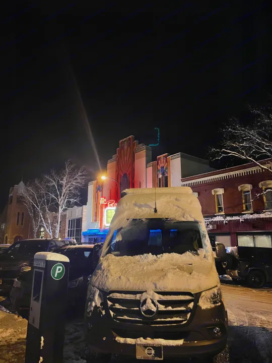A snow-covered Mercedes-Benz RV rental van parked on a winter night street in front of an illuminated Art Deco theater.. StoryTeller Overland Classic Mode 4X4 2022